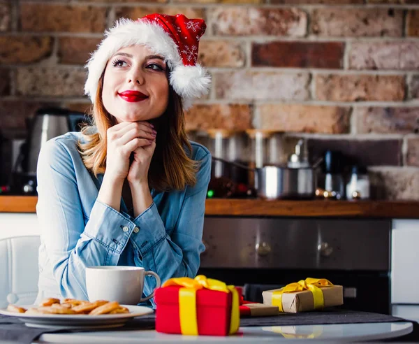 贈り物にキッチンで座っているサンタ クロースの帽子の美しい若い女性 クリスマスの時期 — ストック写真