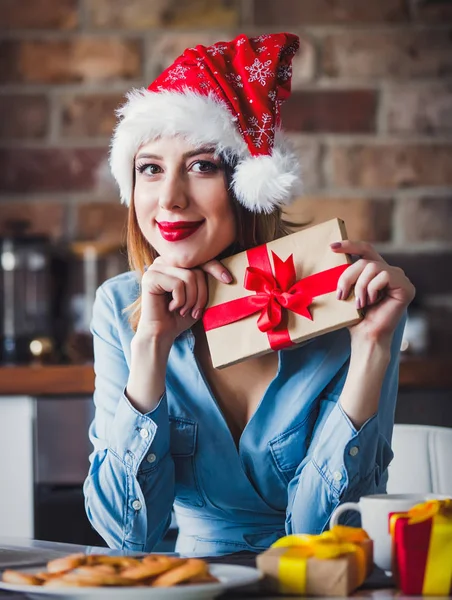 贈り物にキッチンで座っているサンタ クロースの帽子の美しい若い女性 クリスマスの時期 — ストック写真