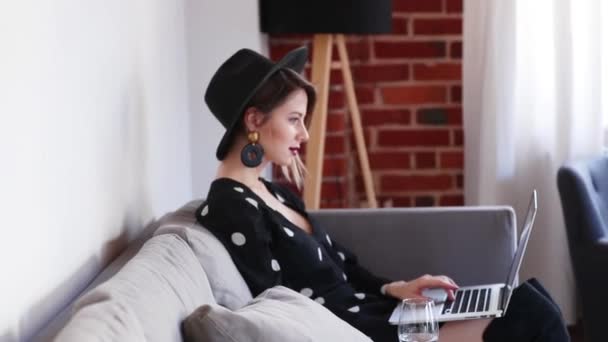 Девушка в черной шляпе и платье сидит на диване с ноутбуком — стоковое видео