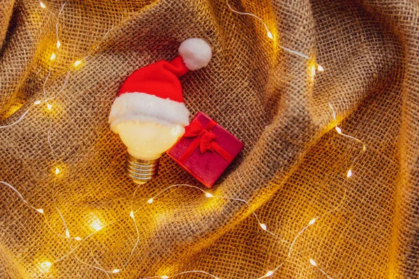 Jüt Arka Plan Üzerinde Noel Şapka Gfit Kutusunda Ampul — Stok fotoğraf