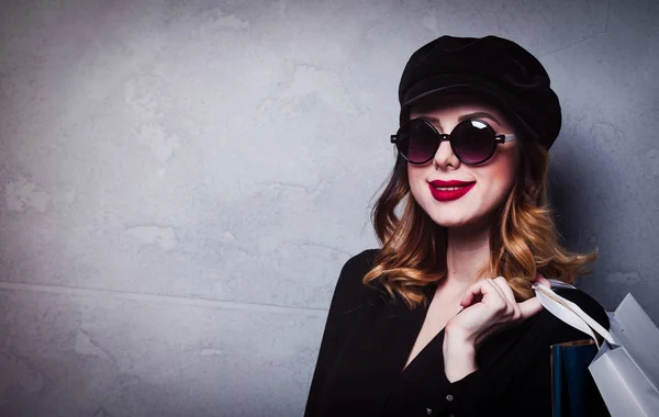 ショッピング バッグや灰色の背景にサングラスと帽子でスタイル赤毛の女の子 — ストック写真