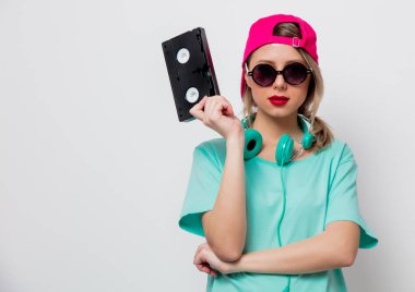 Beautiful young girl in pink cap and blue t-shirt with VHS cassette on white background. clipart