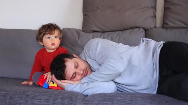 Menino sentado em um sofá perto cansado adormecido pai — Vídeo de Stock