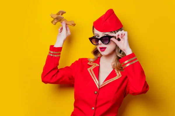 穿着红色制服和与飞机的太阳镜的迷人的复古空姐的肖像 在黄色背景上隔离 — 图库照片