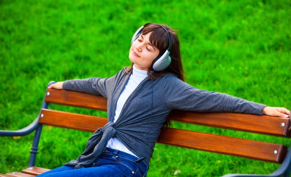 Brunet tjej med hörlurar som sitter på bänken i parken. — Stockfoto