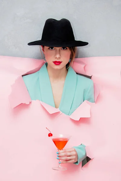 美丽的女人在帽子和蓝色经典夹克与马提尼玻璃看出来的形式撕裂的纸 背景粉红色 — 图库照片