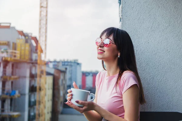 戴着太阳镜的年轻女子, 喝咖啡, 住在阳台上 — 图库照片