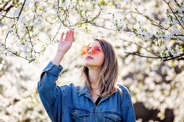 Молодая девушка в джинсовой куртке и солнечных очках стоит рядом с цветком — стоковое фото