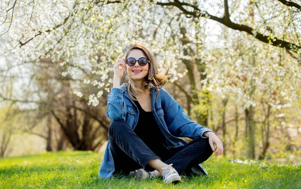 Ung flicka i en jeans jacka och sol glasögon sitter nära en blo — Stockfoto