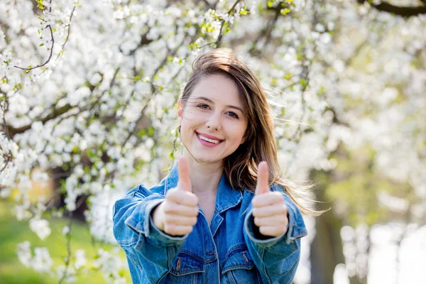 Дівчина в джинсовій куртці стоїть біля квітучого дерева і показує Добре — стокове фото