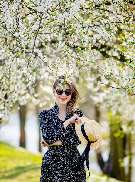 Chica joven en gafas de sol y sombrero permanecer cerca de un árbol con flores —  Fotos de Stock