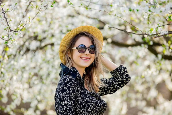 Jong meisje in een zonnebril en hoed verblijf in de buurt van een bloeiende boom — Stockfoto