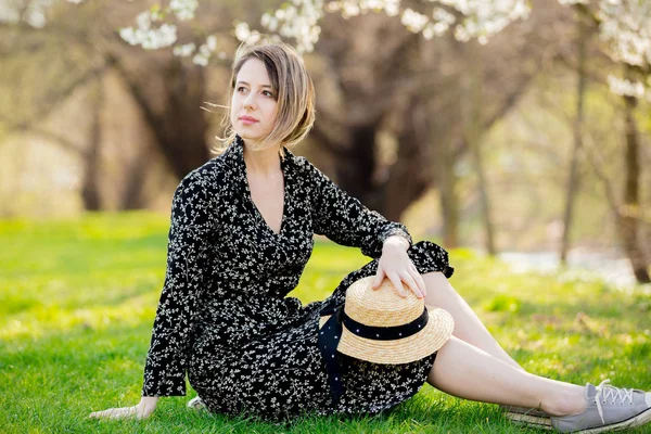 Young girl with hat sitting near a flowering tree in the park. — Stock Photo, Image