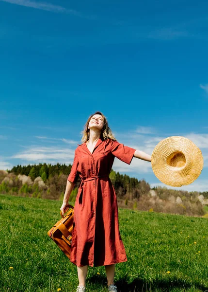 Beautiful girl in red dress and hat with suitcase on meadow — Stock Photo, Image