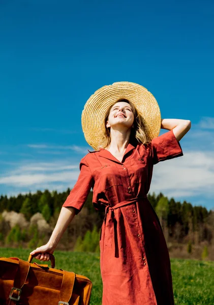 Mooi meisje in rode jurk en muts met koffer op weide — Stockfoto