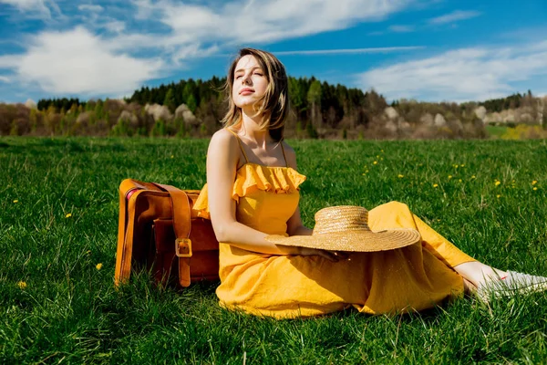 Девушка в жёлтом платье и чемодане, сидящая на горном лугу — стоковое фото