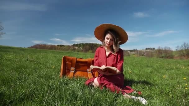 美丽的女孩在红色礼服和帽子与手提箱和书坐在草地上 — 图库视频影像