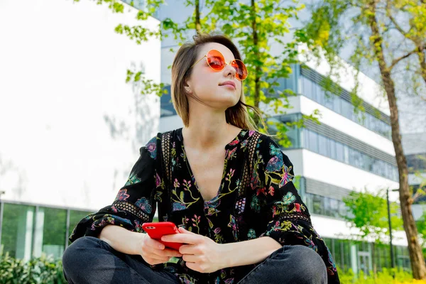 Молодая женщина в солнечных очках с мобильным телефоном сидит на зеленом все — стоковое фото
