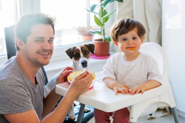 Küçük bir çocuğu kaşıkla ve köpekle besleyen genç bir baba. 