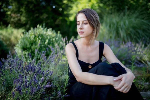 ラベンダーの花の近くに座っている暗いドレスを着た若い女性 — ストック写真
