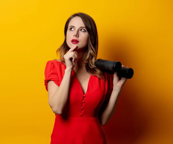 Young woman in red dress with binocular on yellow background — Stock Photo, Image