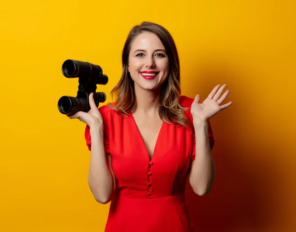 Young woman in red dress with binocular on yellow background — Stockfoto