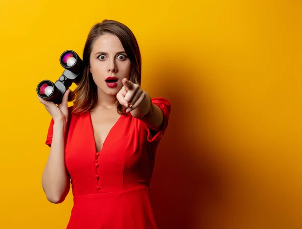 Jovem em vestido vermelho com binocular no fundo amarelo — Fotografia de Stock