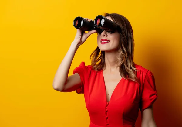 Mujer joven en vestido rojo con binocular sobre fondo amarillo — Foto de Stock