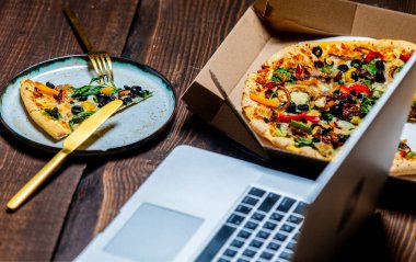 Kartonda zeytin ve peynirli Akdeniz pizzası. Tahta masadaki dizüstü bilgisayarın yanında.