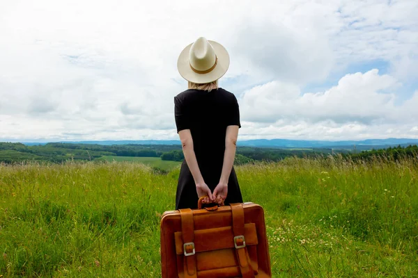 布隆得女人身穿黑衣 提着手提箱站在草地上 背景是群山丛生 — 图库照片