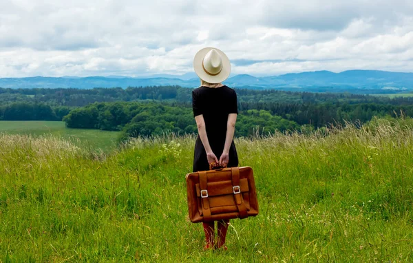 布隆得女人身穿黑衣 提着手提箱站在草地上 背景是群山丛生 — 图库照片