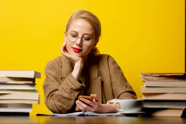 ブロンドの女性は本とテーブルに座って黄色の背景に携帯電話を使って — ストック写真