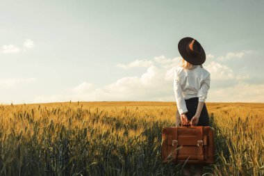 Gün batımında buğday tarlasında bavullu güzel sarışın kız.