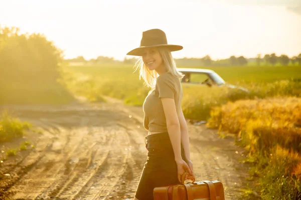 美しいですブロンド女の子とともにスーツケースで田舎道で日没時間 — ストック写真