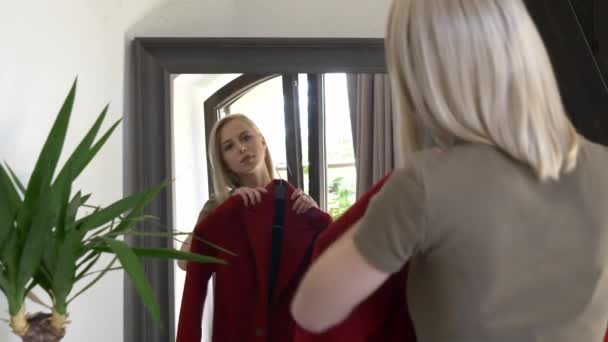 ブロンドの女の子が家でミラーで秋のシーズンのために赤いコートを着ている — ストック動画