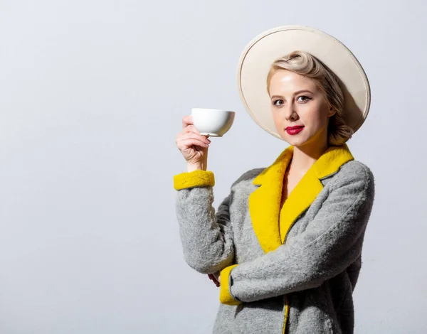 ブロンド女の子でグレーコートとヴィンテージヘアドレスともにコーヒーのカップの白の背景 — ストック写真