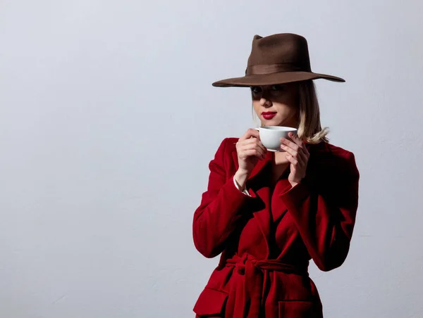 赤いコートのブロンドの女の子とグレーの背景にコーヒーのカップとヴィンテージの帽子 ノアスタイルの画像 — ストック写真
