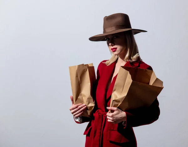 赤いコートのブロンドの女の子とグレーの背景に紙のスーパーマーケットバッグとヴィンテージの帽子 ノアスタイルの画像 — ストック写真