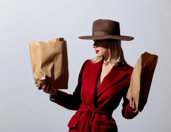 赤いコートのブロンドの女の子とグレーの背景に紙のスーパーマーケットバッグとヴィンテージの帽子 ノアスタイルの画像 — ストック写真