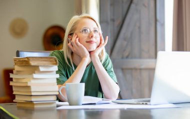 Gözlüklü sarışın kız defterle çalışıyor ve evde serbest çalışan biri olarak kahve içiyor.