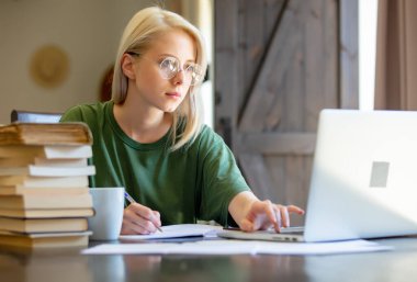 Gözlüklü sarışın kız defterle çalışıyor ve evde serbest çalışan biri olarak kahve içiyor.