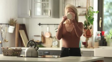 Sarışın kadın mutfakta elinde bir fincan kahveyle duruyor.