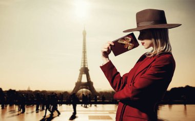 Kırmızı ceketli sarışın kız Eyfel kulesinin arkasında hediye kutusu var. 