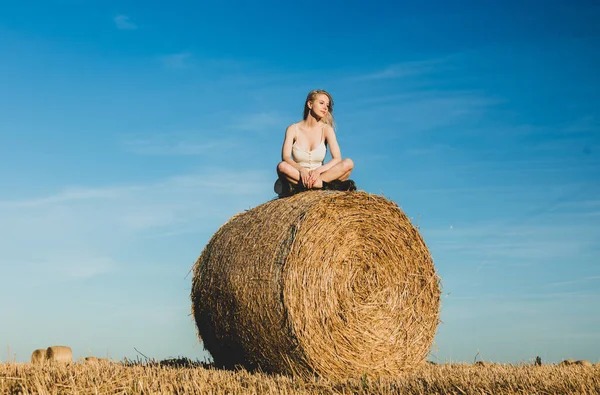 ブロンドの女の子は日没の時間に野外で転がされた干し草の上に座っている — ストック写真