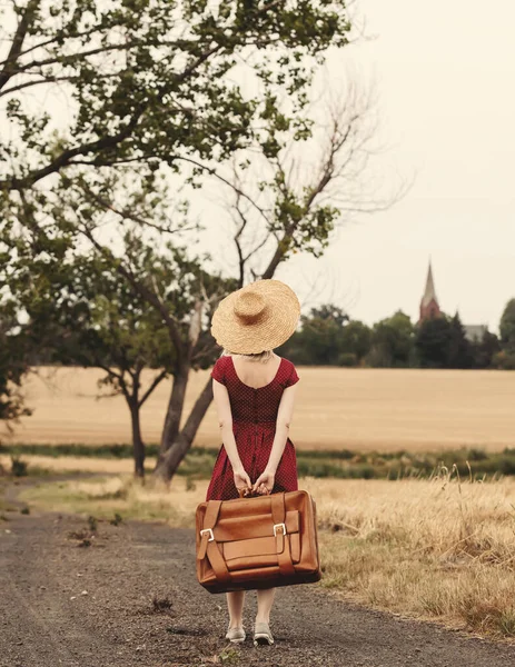 blonde in red dress with a suitcase on a rural road before the rain