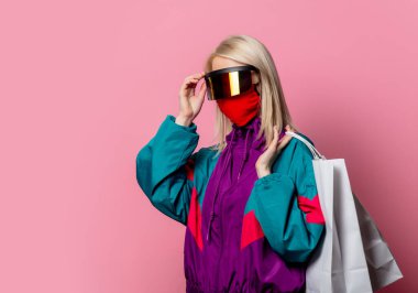 80 'lerin kıyafetleri ve maskesiyle pembe arka planda alışveriş torbaları olan sarışın.