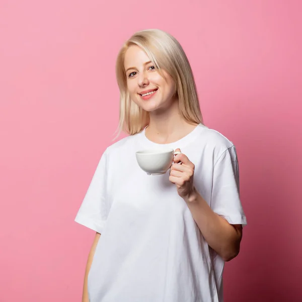 ブロンド女性でホワイトMoch Tシャツともにコーヒーのカップのピンクの背景 — ストック写真