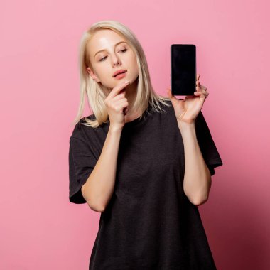 Pembe arka planda siyah tişörtlü ve cep telefonlu sarışın kadın.