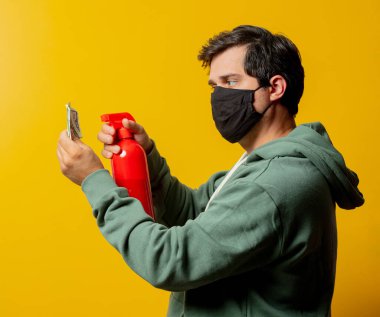 Yüz maskeli adam dezenfeksiyon spreyi ve sarı arka planda para tutuyor.