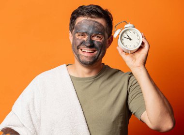 Yüzünde kozmetik maske ve turuncu arka planda çalar saati olan komik bir adam.